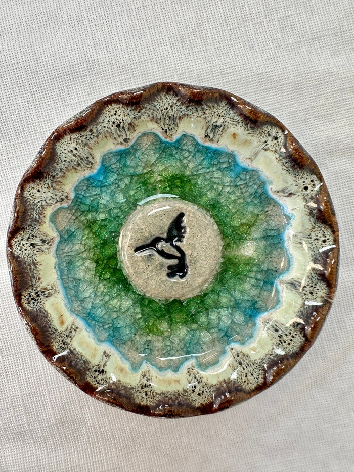 Circular ceramic hummingbird trinket tray with a beige center and a black hummingbird silhouette surrounded by blue to green crackle glaze