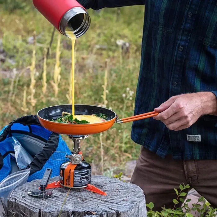 Lifestyle image of a man pouring beaten egg into a Jetboil Summit Skillet whilst being heated by a camping gas stove