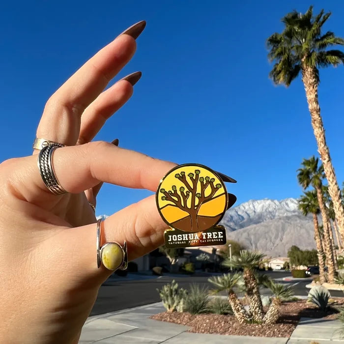 Close up of a hand holding a Joshua Tree enamel pin against a desert backdrop with palm trees and blue sky.