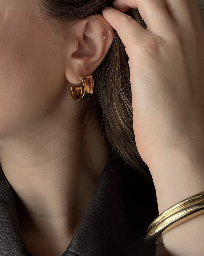 A close-up of a person with light skin wearing a gold hoop earring and the Leila Dome Cuff Bracelet by Mod + Jo, with their hand touching their hair while dressed in a dark, textured top.