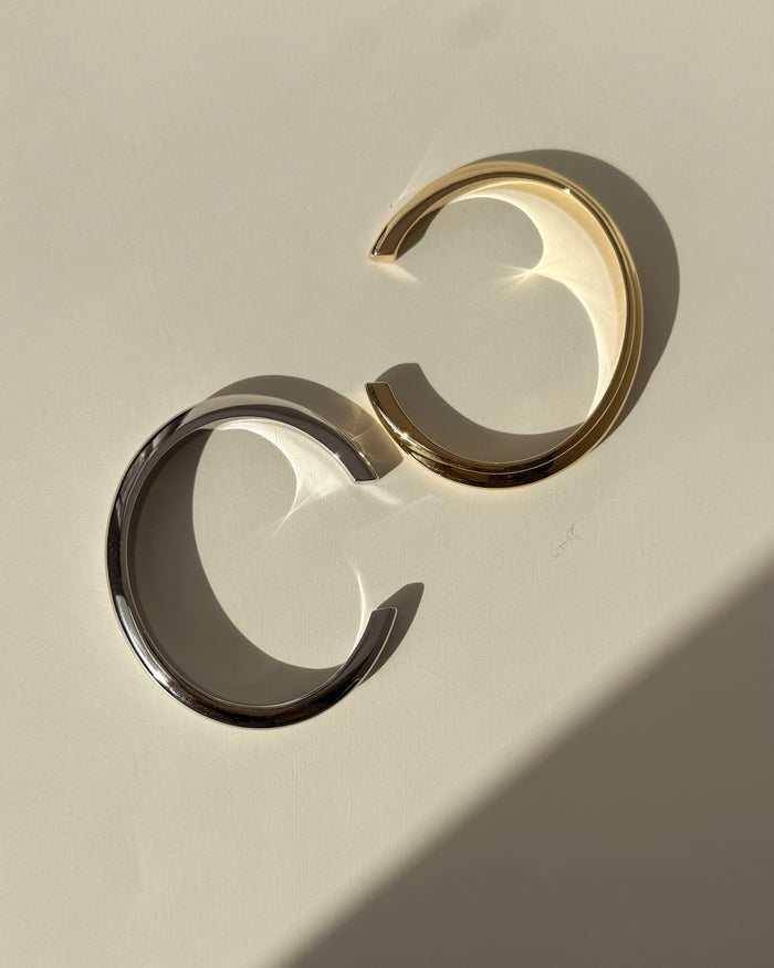 Two Leila Dome Cuff Bracelets by Mod + Jo—one gold plated, one silver—rest on a light surface. Gentle shadows and reflections highlight their elegant design, ideal for any 14k gold jewelry collection.