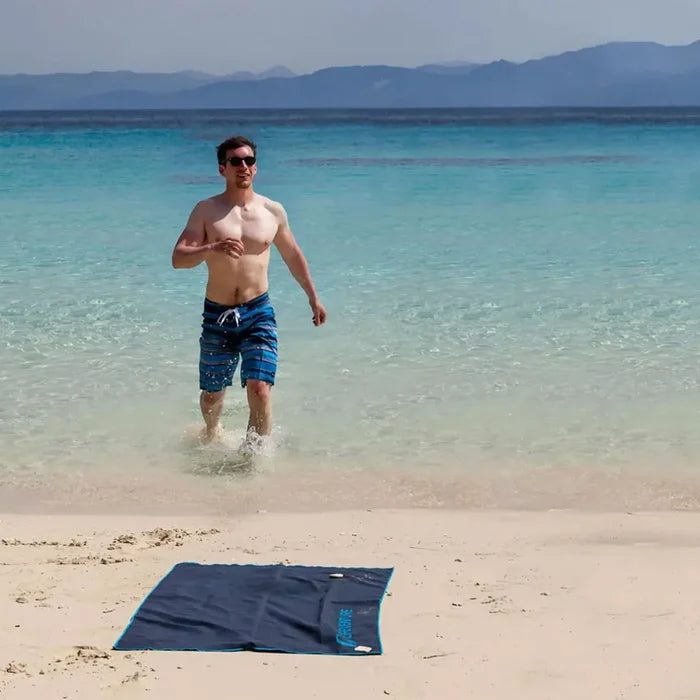Lifestyle image of a male swimmer walking out of the sea on to the beach where his Lifeventure SoftFibre recycled travel towel in grey is laid out.
