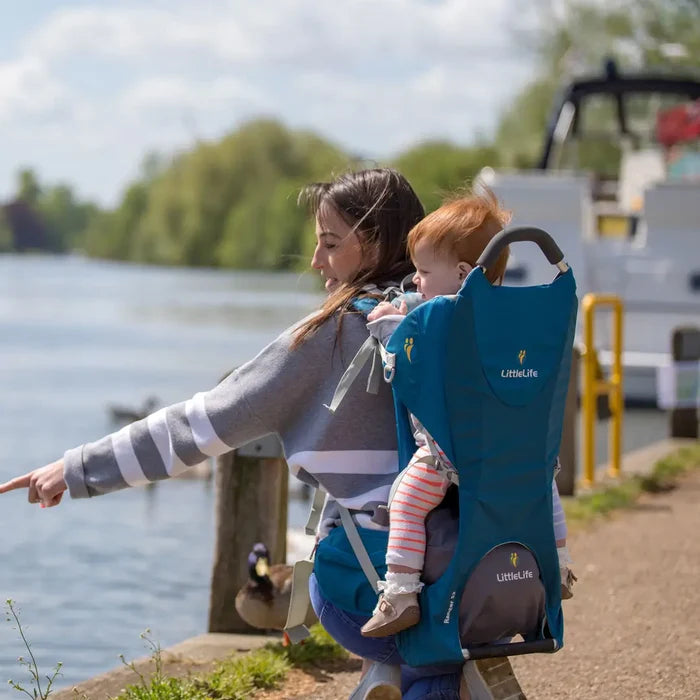 Lifestyle image of a woman wearing a Littlelife Ranger S2 child carrier backpack in blue colour with her child in the back whilst walking along a riverbank