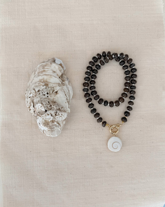 The Mod + Jo Macey Gemstone Necklace, featuring dark round beads, a white spiral shell pendant, and genuine gemstones, is displayed beside a large oyster shell on a light beige fabric background.