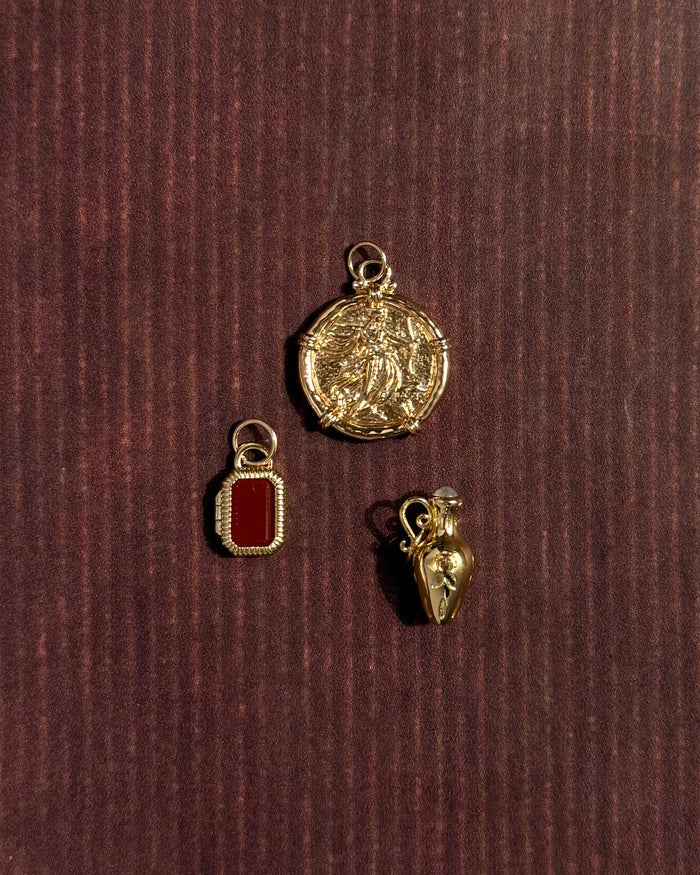 Three 14k gold-plated charms from Mod + Jo rest on a dark, textured surface: a round charm with an intricate design, the Mabel Rectangle Birthstone Locket Charm with a red center, and a heart-shaped charm featuring a small loop.