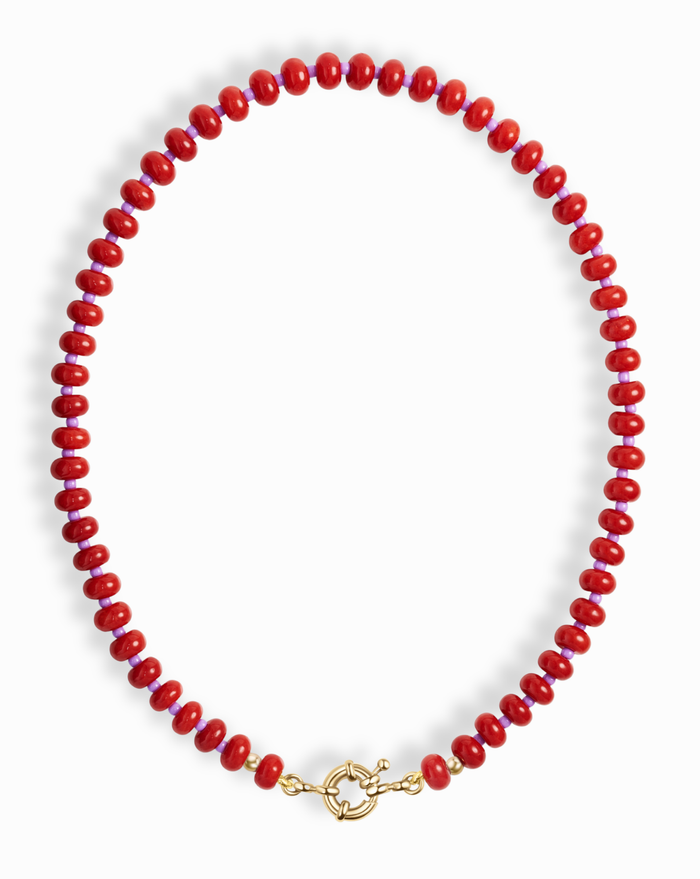 The Macey Gemstone Necklace by Mod + Jo features round, glossy red beads and a gold toggle clasp, artfully displayed in a circular shape on a white background.