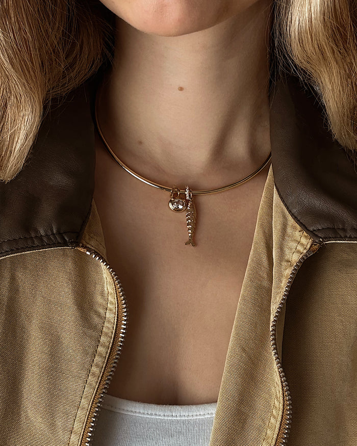 A woman in a brown and tan jacket with a white top wears a gold choker necklace featuring two pendants: a small fish skeleton and the Mod + Jo Marla Sapphire Bezel Charm.