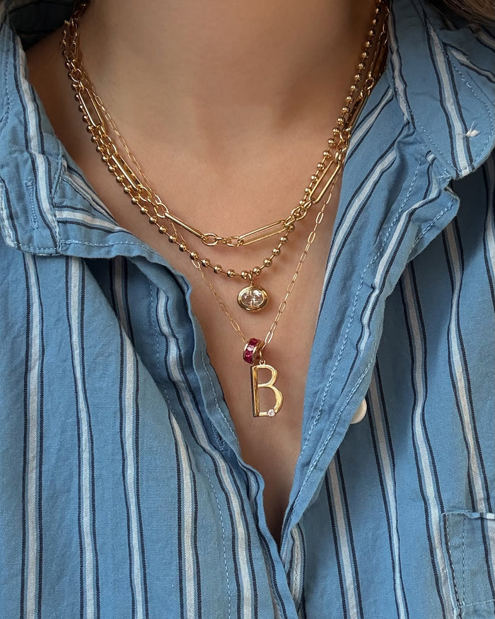 A person wears a blue and white striped shirt and three layered gold necklaces, including Mod + Jo's Marla Sapphire Bezel Charm and a pearl pendant.