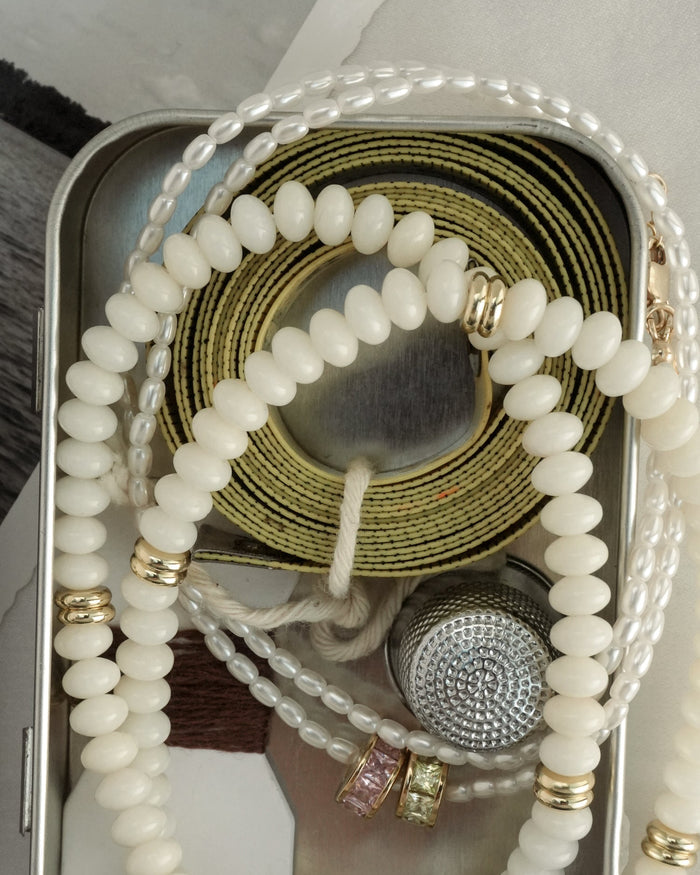 A tin box with white beaded necklaces, the Merit Gemstone Necklace by Mod + Jo, a yellow measuring tape, a silver textured thimble, colorful stone rings, and a partially visible black-and-white photo underneath.