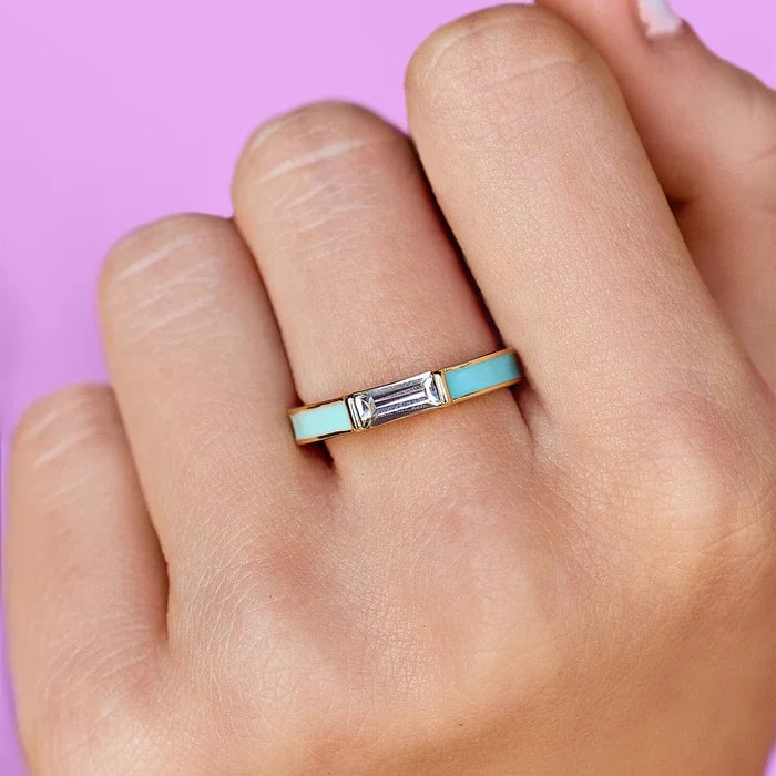 Turquoise enamel two-tone ring with a rectangular baguette gemstone worn on a finger against a pink background.