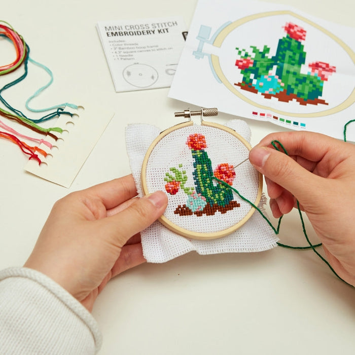 Hands stitching the cactus pattern in a small embroidery hoop with colorful threads and instructional sheets nearby.