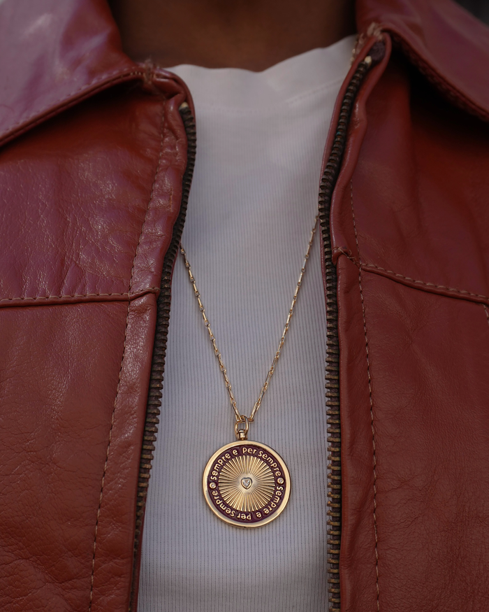 Gold necklace with a circular red enamel pendant with the phrase Sempre e per sempre worn by a person in a red leather jacket.