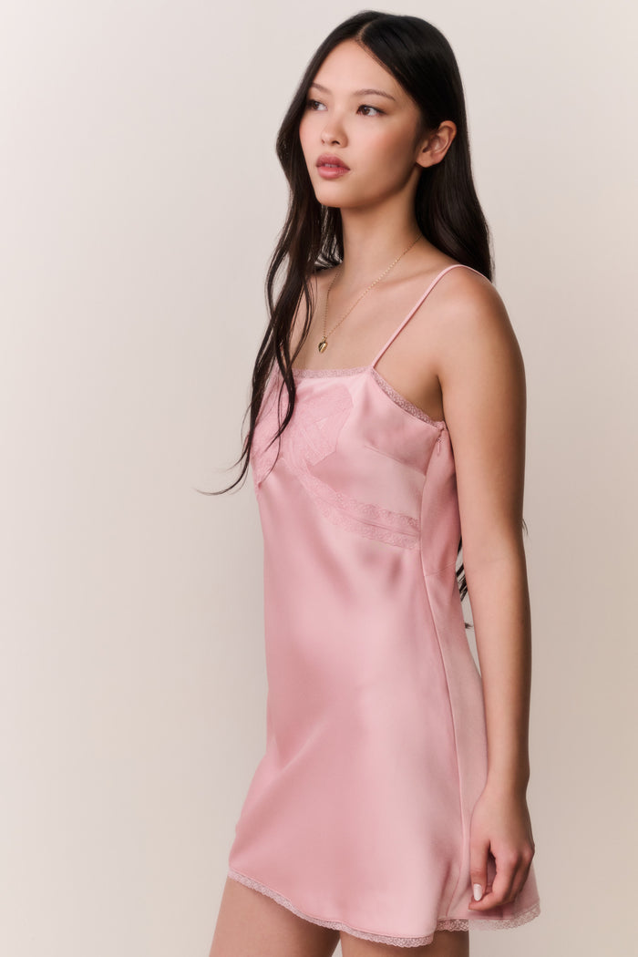 Woman wearing a pink satin slip dress with bow design against a plain background