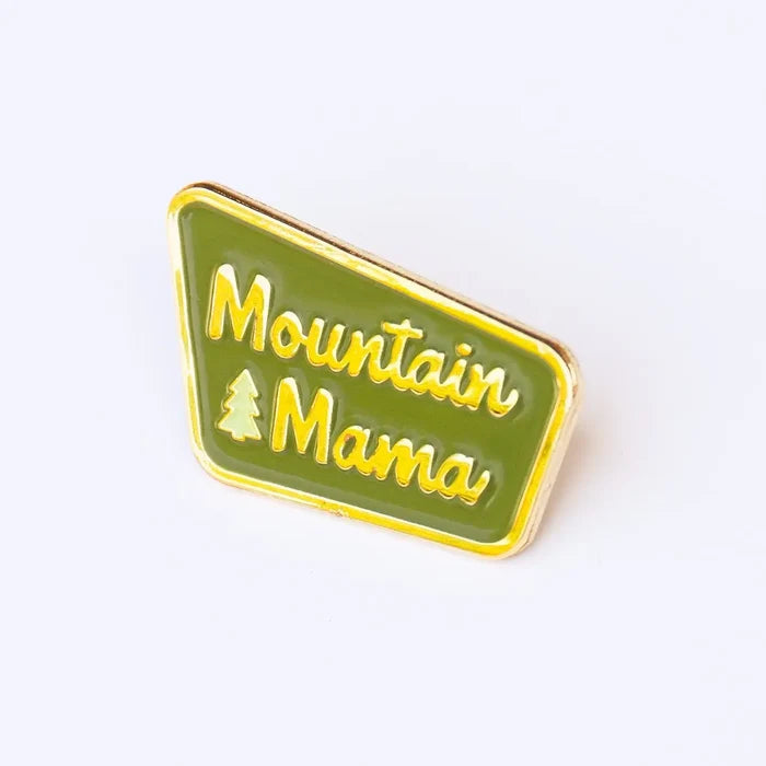 Close up of Mountain Mama enamel pin with green field gold lettering and tree icon