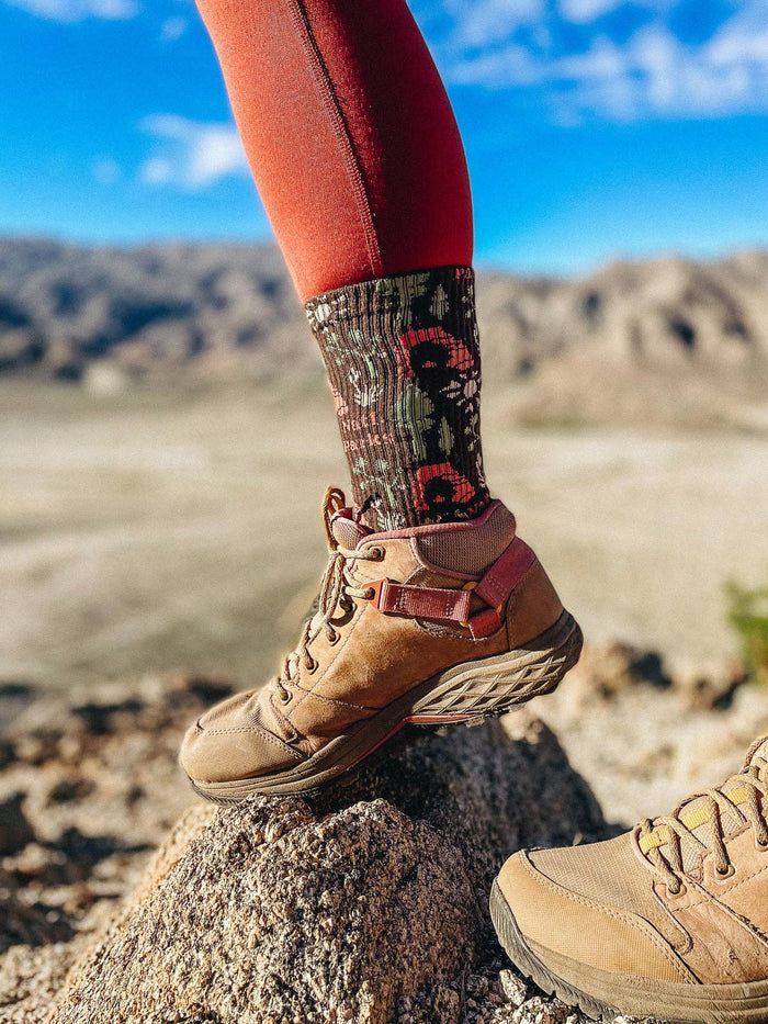 Close up of a hiker wearing National Park Roundup Socks in red with a floral pattern and a tan hiking boot on a rock