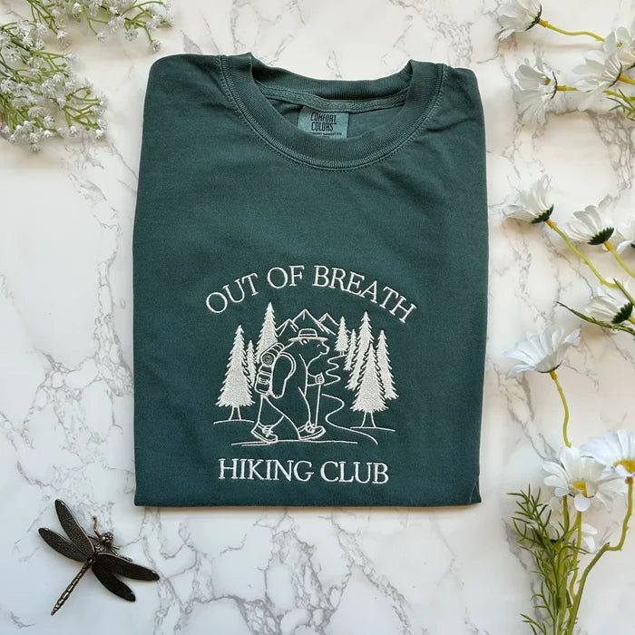 Teal hiking club tee with white embroidery of a hiker among trees and mountains and the text Out of Breath Hiking Club