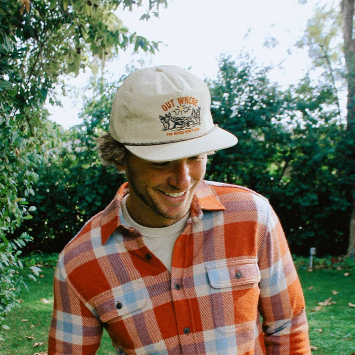 Beige cap with OUT WHERE THE BUSES DON'T RUN embroidery worn by a man in an orange plaid shirt outdoors