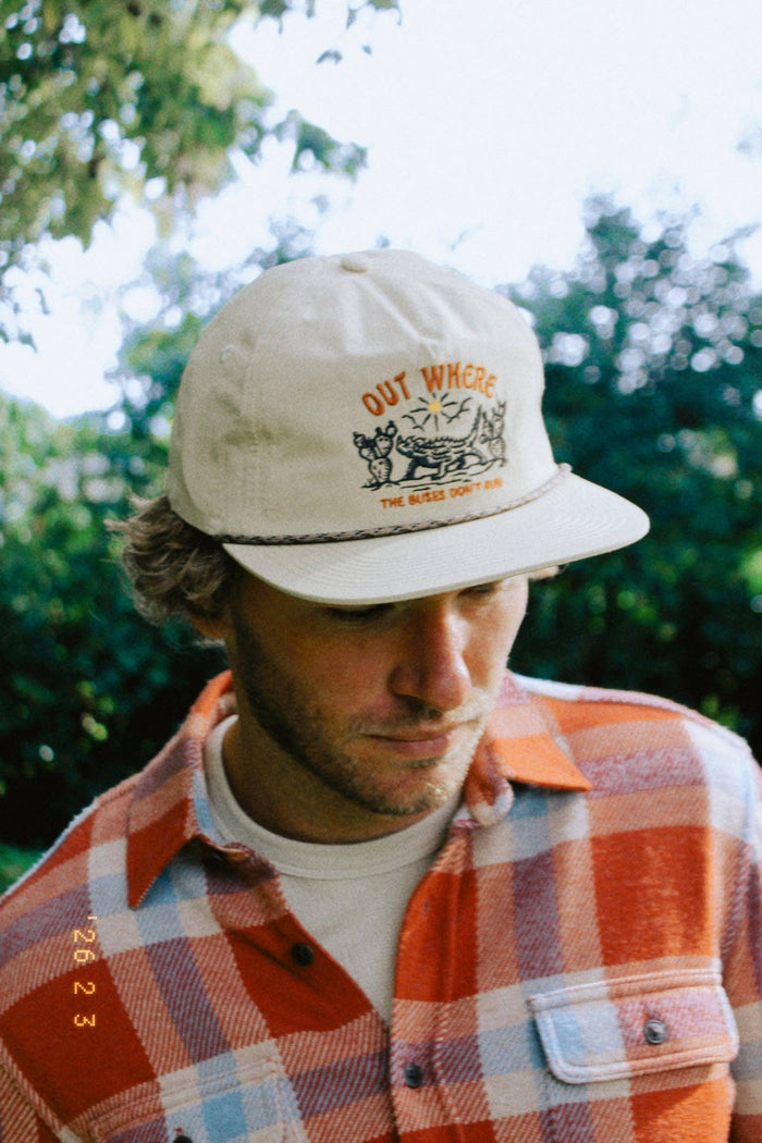 Beige cap with OUT WHERE THE BUSES DON'T RUN embroidery and illustrated scene worn by a man in an orange plaid shirt