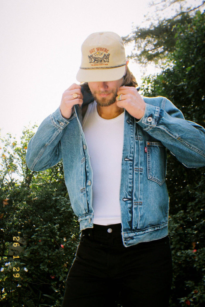 Man wearing a beige Out Where The Buses Don't Run hat with braided band and a denim jacket outdoors.