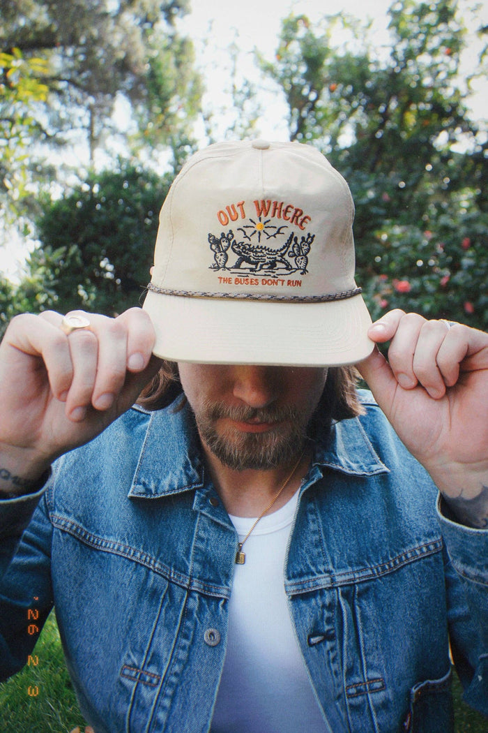 Close up of man wearing the beige Out Where The Buses Don't Run hat, adjusting his denim jacket outdoors.