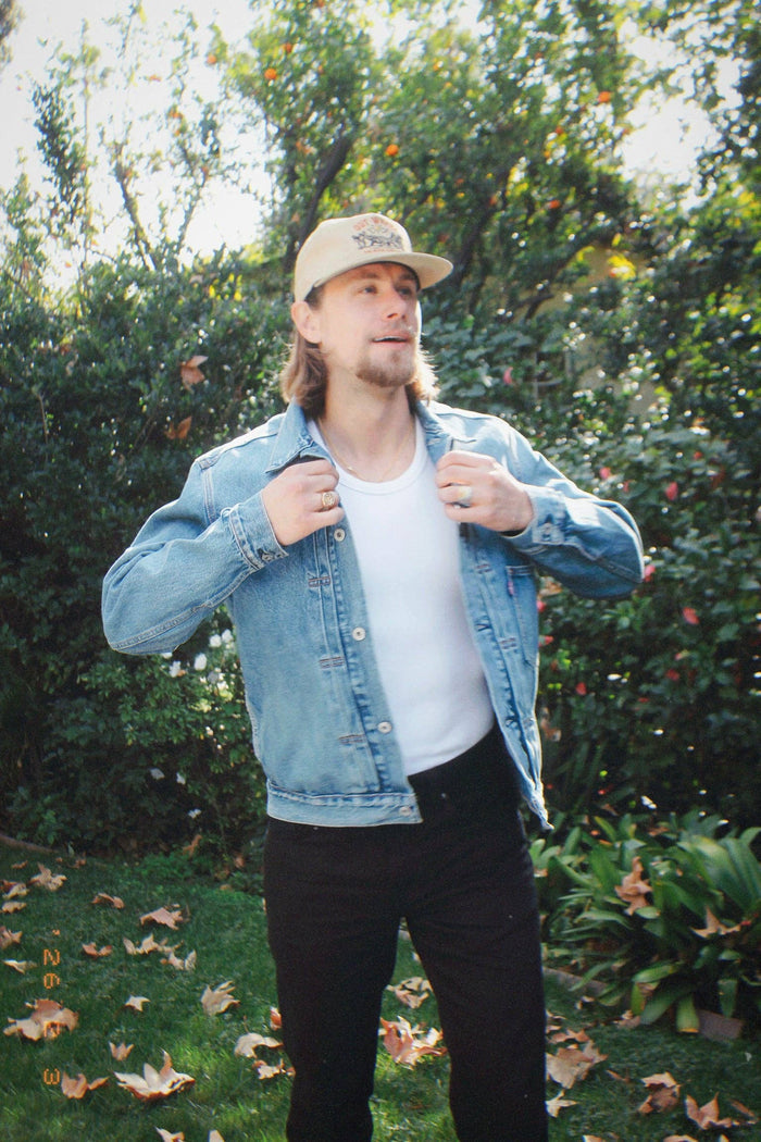 Full body of a man in a denim jacket and white tee wearing the beige logo hat outdoors in a garden.