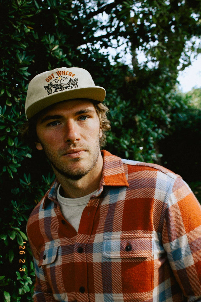 Beige cap with Out Where The Buses Don't Run embroidery worn by a man in a plaid shirt outdoors.