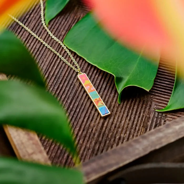 Paradise Island Reversible Bar Necklace vertical with a colorful mosaic pendant on a gold chain resting on wood and tropical leaves.