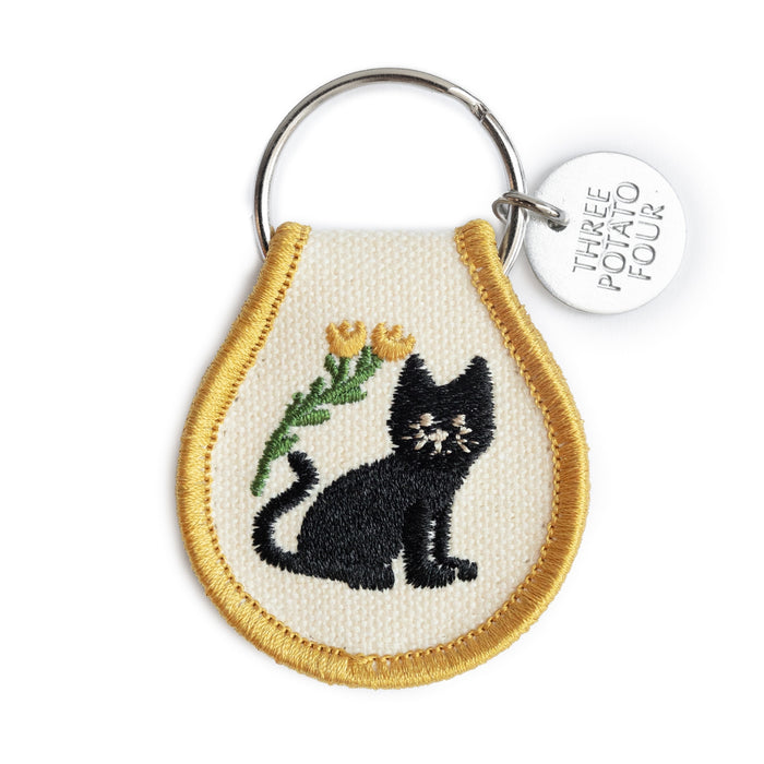Patch keychain with a black cat embroidered on cream fabric bordered in gold, attached to a silver ring and round metal tag.