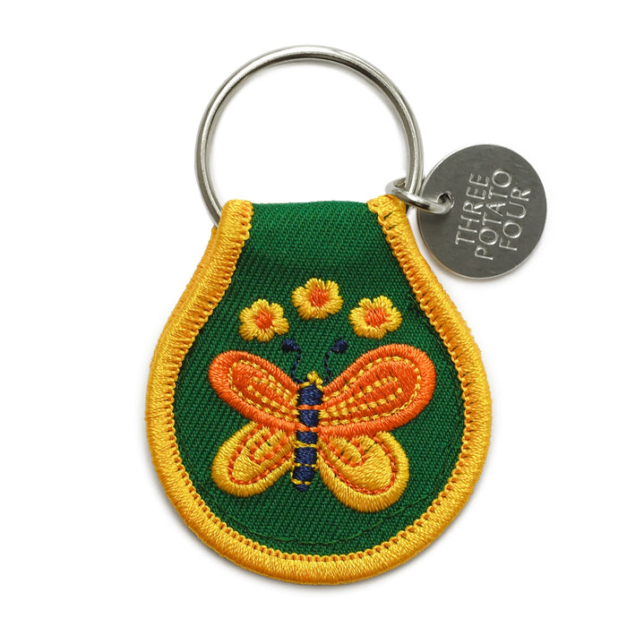 Patch keychain with green fabric and a yellow border featuring an orange and yellow butterfly embroidery, attached to a silver ring and round metal tag.