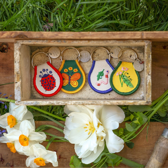 Five embroidered patch keychains displayed in a rustic wooden box with metal rings and round tags