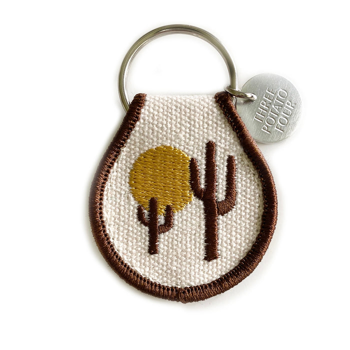 Teardrop shaped embroidered desert patch keychain with sun and cacti on cream fabric and brown trim.