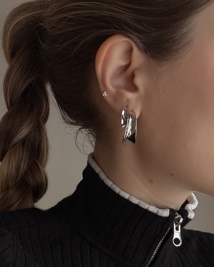 Close-up of a person with braided brown hair wearing a black ribbed zip-up top and Mod + Jo's Grande Valen Tapered Hoops—large, shiny, rhodium-plated earrings—in one ear. The image shows part of their face and neck.