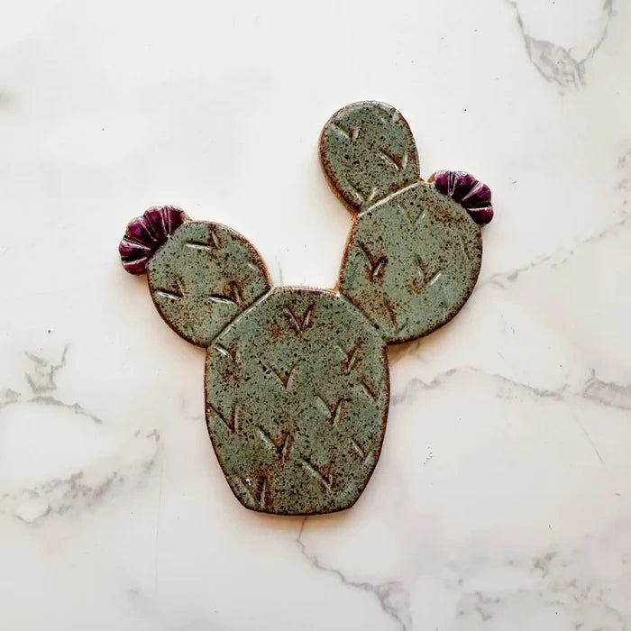 Prickly pear cactus ceramic magnet with three green speckled pads and purple flowers on a light surface.
