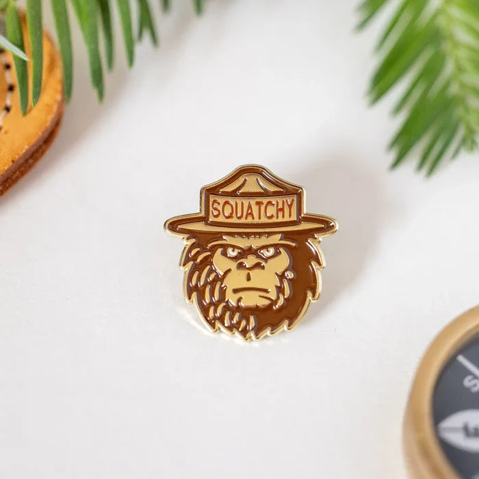 Ranger Squatchy enamel pin featuring a Sasquatch wearing a ranger hat on a white surface with green plant fronds