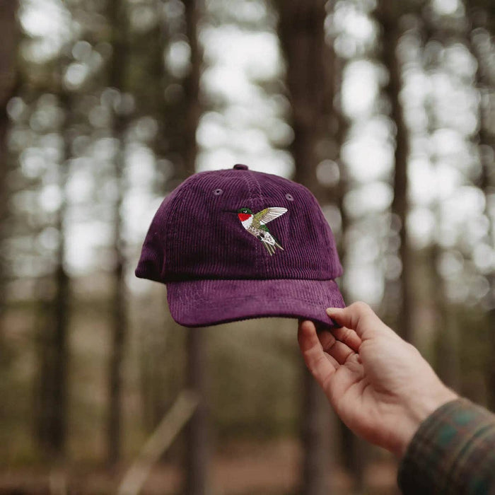 Purple corduroy hat with hummingbird embroidery held outdoors in a forest