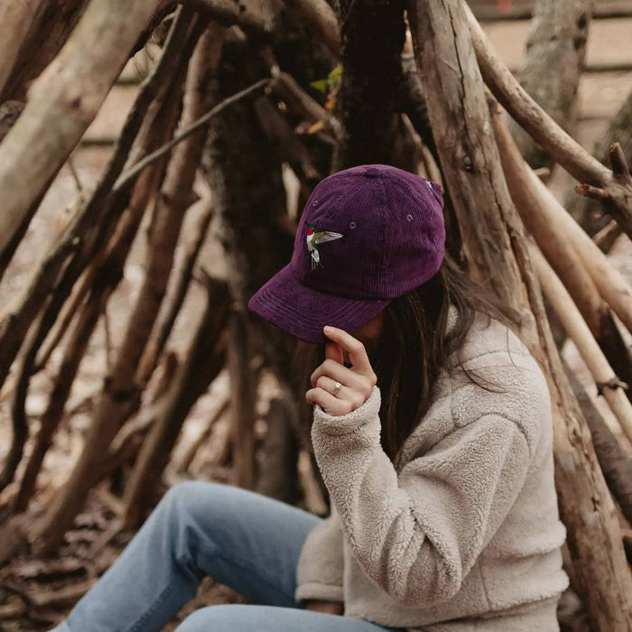Person wearing a purple corduroy hummingbird hat with embroidery among wooden branches.