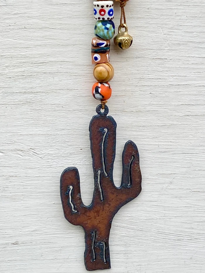 Colorful beaded strand with a rustic cactus metal pendant and a brass bell against a light wooden background.