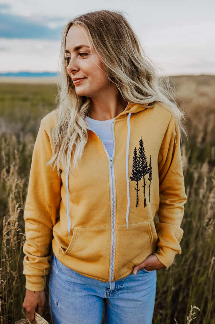 Mustard Rustic Three Tree zip hoodie worn by a person in a field, showcasing three tree graphic on the chest.