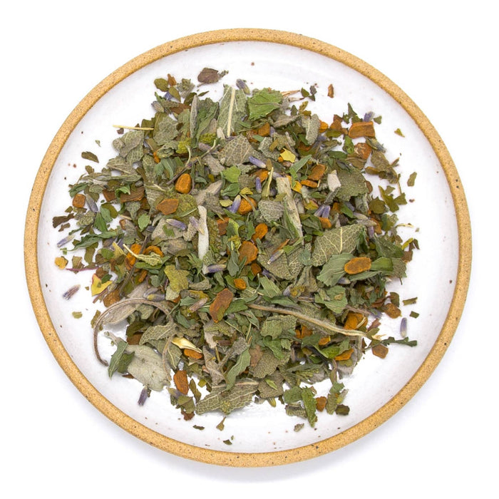 Sage and Friends Herbal Tea loose blend with dried green leaves orange chunks and purple flowers on a round white plate.