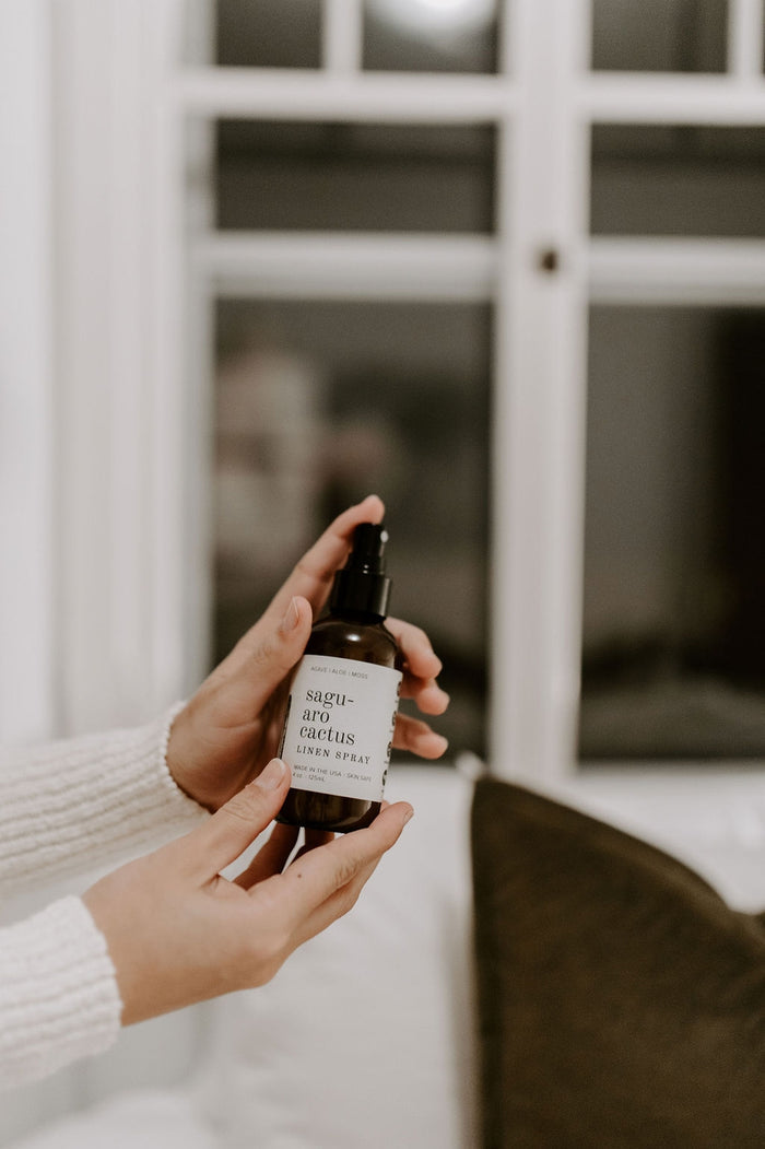 Hands hold Saguaro Cactus Linen Spray bottle in a softly lit room by a window