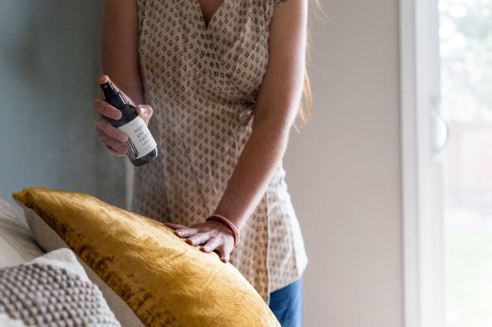 Person in a patterned sleeveless dress holds Saguaro Cactus Linen Spray beside a gold pillow on a bed