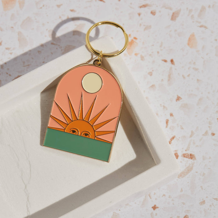 Setting Sun enamel keychain with a peach arch, orange sun rays and a pale circle above, attached to a gold ring.