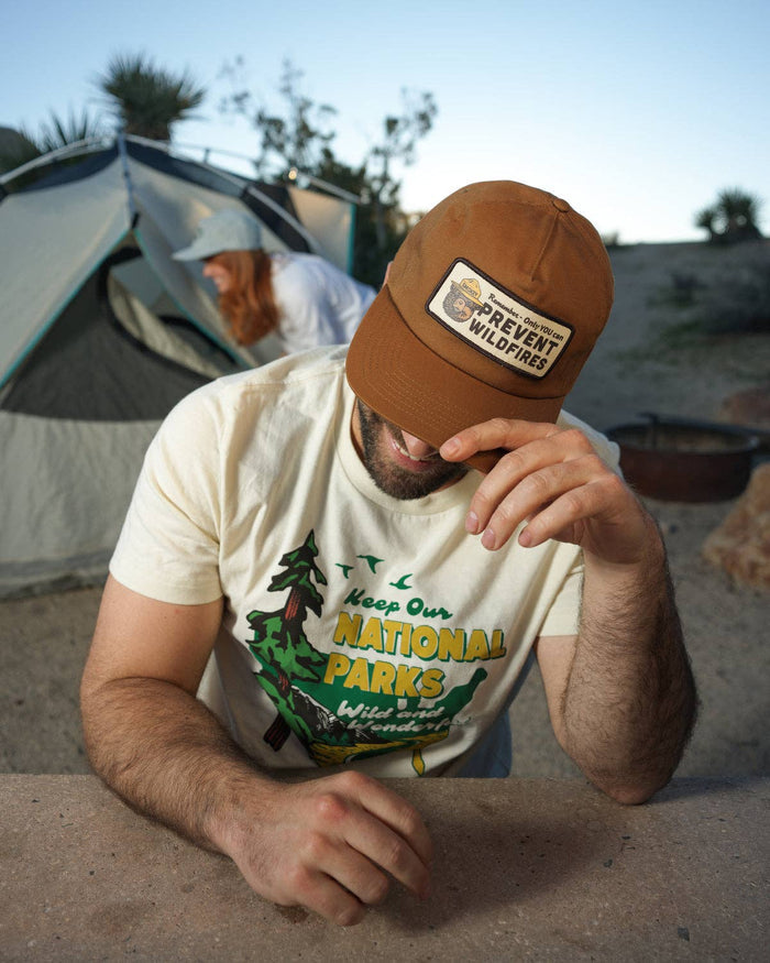 Man wearing a brown Smokey's Reminder five-panel trucker hat at a campsite with a tent in the background