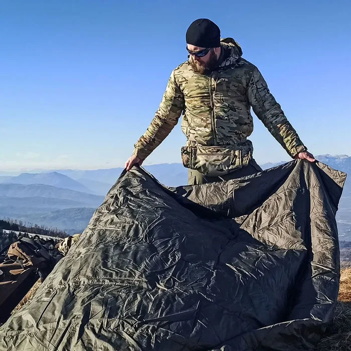 Lifestyle image of a man laying out a Snugpak Jungle Blanket XL in olive on the ground whilst outdoors backpacking