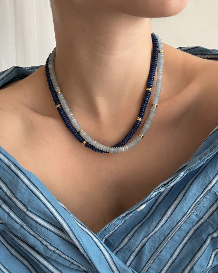 Someone wears the Sonia Gemstone Necklace by Mod + Jo, featuring blue and gray beads with gold accents, paired with a blue and white off-shoulder striped top.