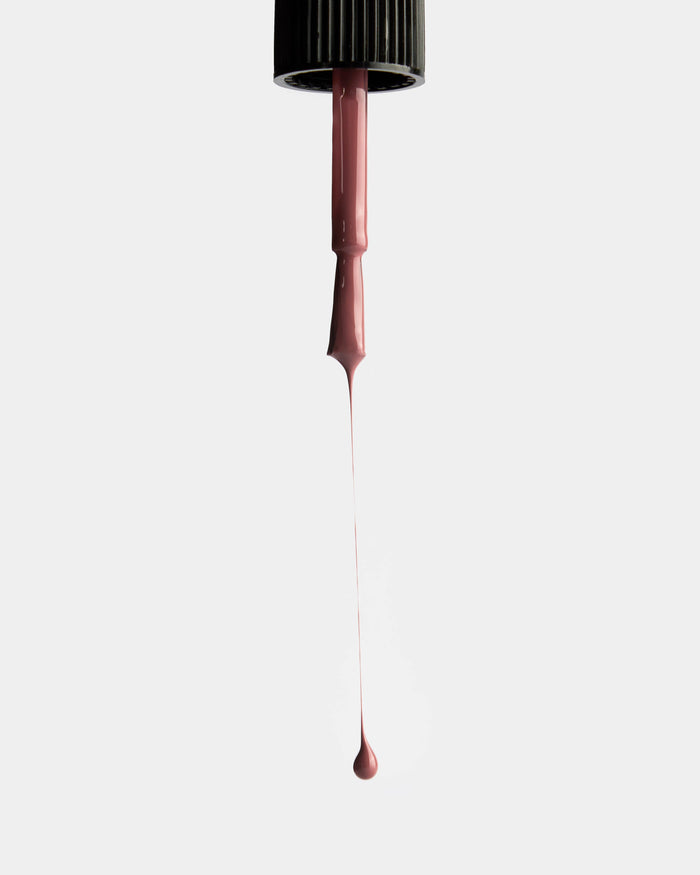 Dusty rose nail polish being dripped from a nail polish application brush against a white background