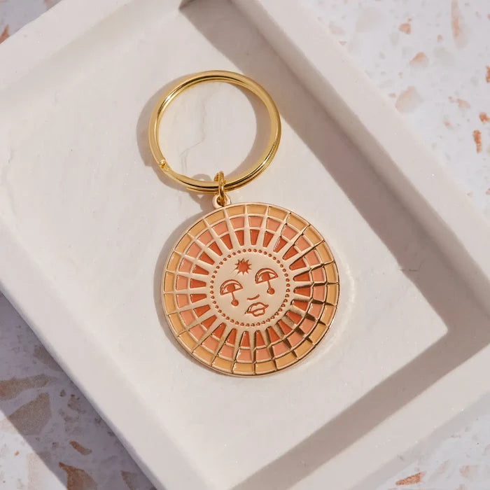 Round sun enamel keychain with peach and pink rays and a smiling face attached to a gold ring.