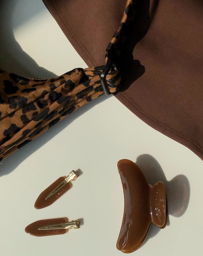 The SuperClip Hair Claw, a brown garment, a leopard-print fabric bag, and two brown and gold hair clips are arranged on a white surface in soft sunlight.