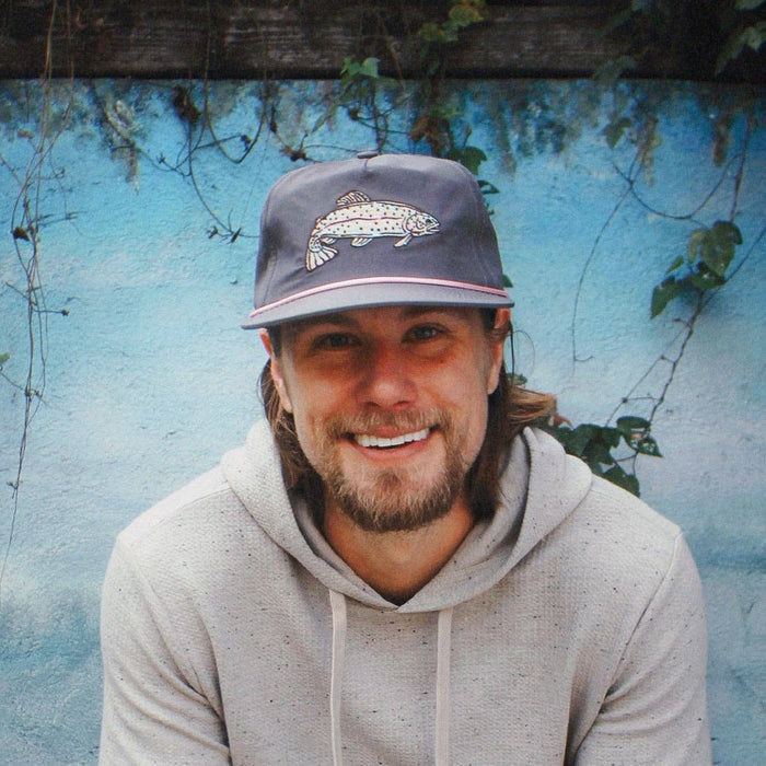 Man wearing The Trout Hat gray cap with fish embroidery in front of a blue wall.
