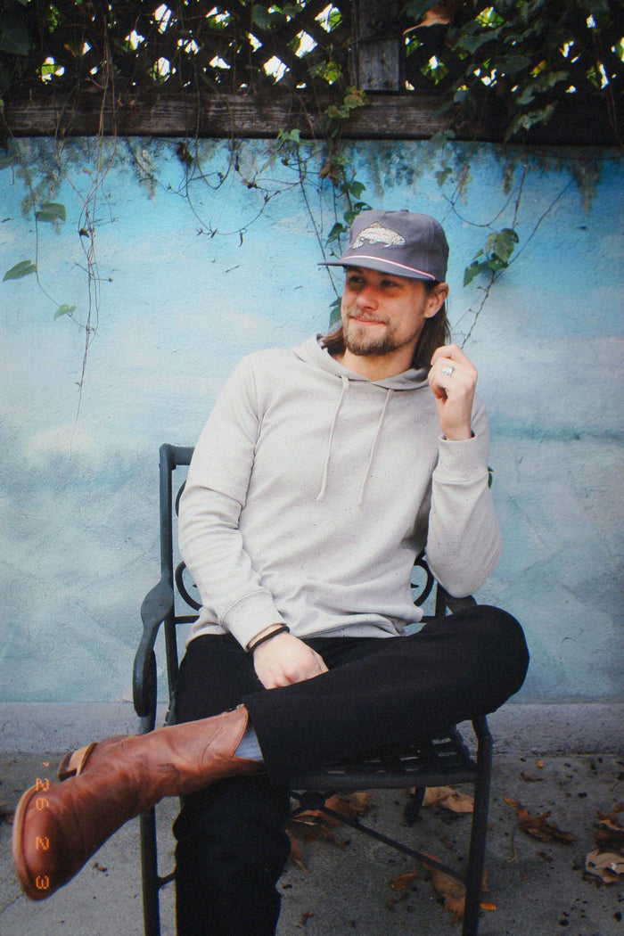 Man wearing The Trout Hat and a light gray hoodie sits on a metal chair outdoors with a blue wall behind and autumn leaves on the ground.